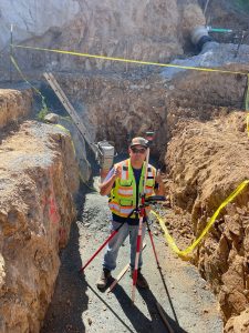 Crew Chief Jared Nielsen surveys an excavation site with a determined expression, holding surveying equipment against a backdrop of heavy machinery and a partially dug foundation. The image showcases professionalism, precision, and leadership in construction management.