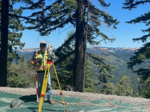 Crew Chief Jared Nielsen surveys a project in the Santa Cruz Mountains, surrounded by towering trees and rugged terrain. Holding a surveying instrument, he demonstrates expertise and adaptability in a challenging outdoor environment.