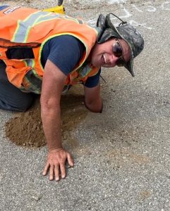 Crew Chief Miles Wadsworth is captured mid-action, digging with determination in a rugged outdoor setting. The scene exudes grit, focus, and a sense of adventure, symbolizing the pursuit of excellence in challenging environments.